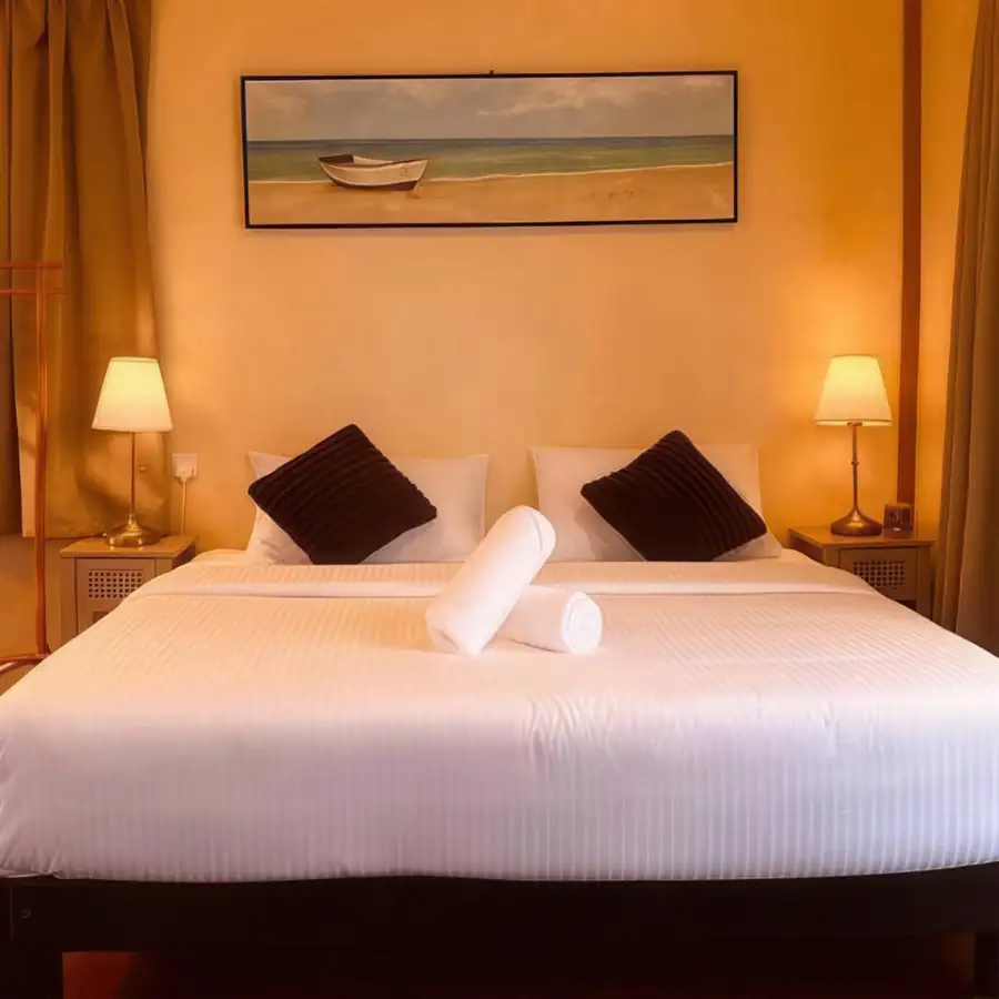 A warm bedroom at Coco Seaweed Resort in the Celebes Sea highlighting a large bed, decorative brown pillows, glowing bedside lamps, boat wall art, polished teak wood floors, and aircon.